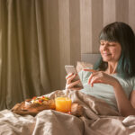 Gesundes Frühstück im Bett. Eine junge Frau im Schlafanzug hält ein Telefon, auf den Knien steht ein Tablett mit einer Schüssel Müsli, Obst und ein Glas mit Orangensaft.