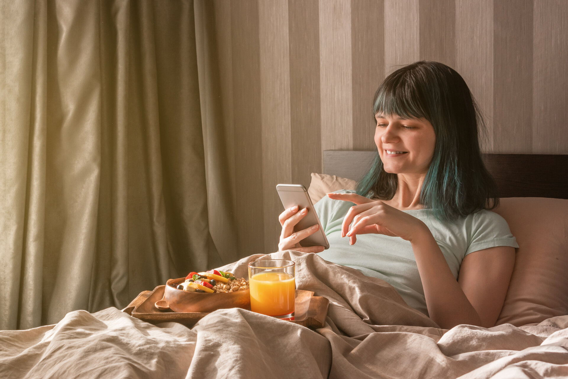 Gesundes Frühstück im Bett. Eine junge Frau im Schlafanzug hält ein Telefon, auf den Knien steht ein Tablett mit einer Schüssel Müsli, Obst und ein Glas mit Orangensaft.