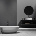 Dunkles Badezimmer mit weißer Badewanne auf grauem Betonboden, einem Waschbecken mit Waschtischunterschrank einem runden Spiegel und Handtuchhalterleiter.
